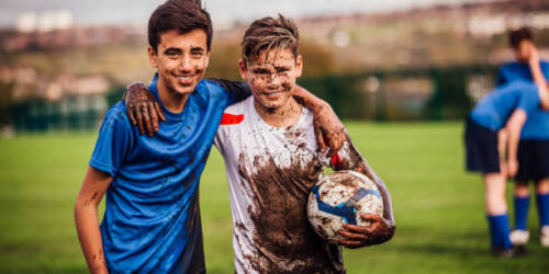 Teenage boys looking at the camera smiling after their soccer game. Both covered in mud. Other players can be seen blurred in the background.